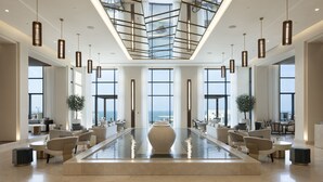 A spacious lobby with a central water feature, large windows offering views, and modern lighting fixtures. - Jumeirah Gulf of Bahrain (Zallaq)