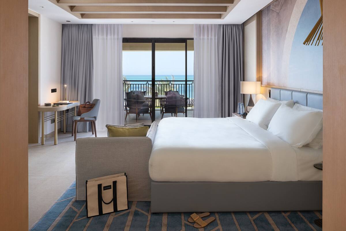 A hotel room with a large bed, a desk with a chair, a small sofa, a balcony with chairs, and a view of the sea.