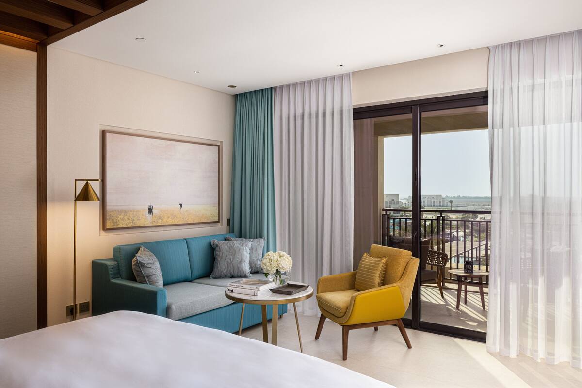A modern hotel room with a bed, a sofa, a yellow armchair, a small table with a vase of flowers, and a balcony with a view.