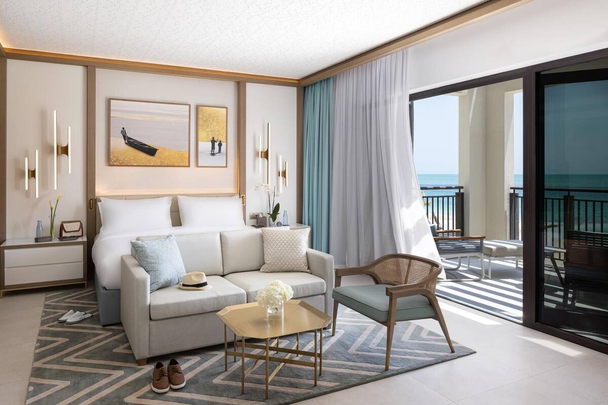 A modern hotel room with a sofa, armchair, and a balcony with a view of the ocean.