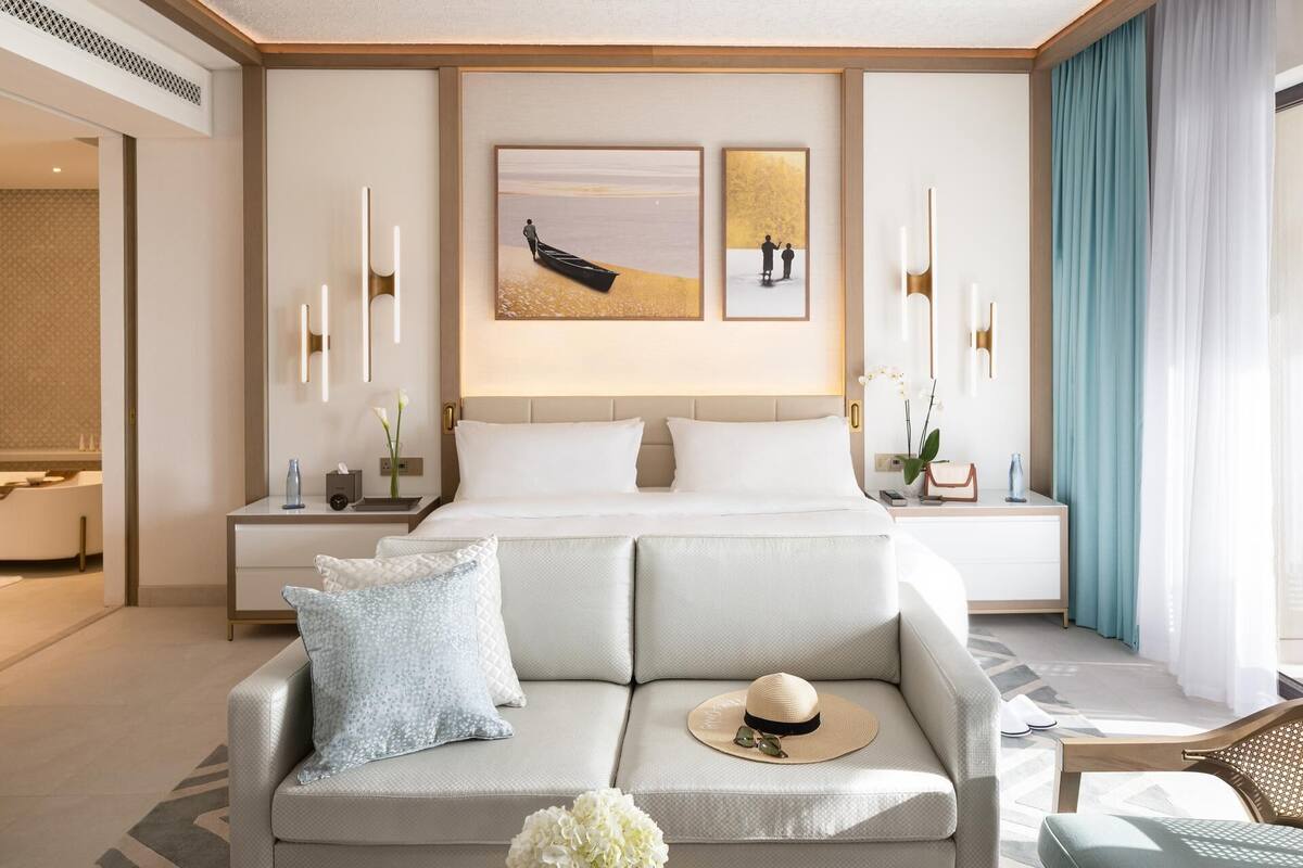 A modern hotel room with a large bed, a sofa, and a desk. There are two framed pictures on the wall, a vase of flowers on the sofa, and a hat on the armrest.