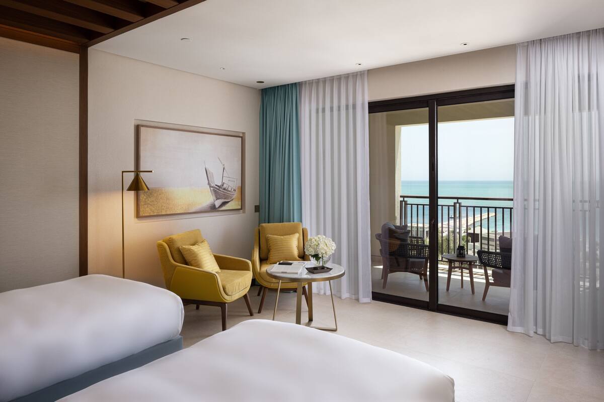 A hotel room with two beds, a small table with a vase, yellow chairs, and a balcony with a view of the sea.