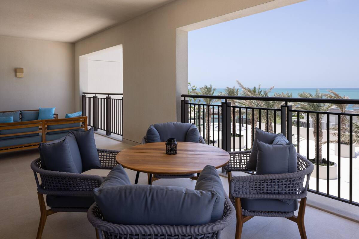 A balcony with wicker furniture, a round wooden table, and a view of the beach and ocean.