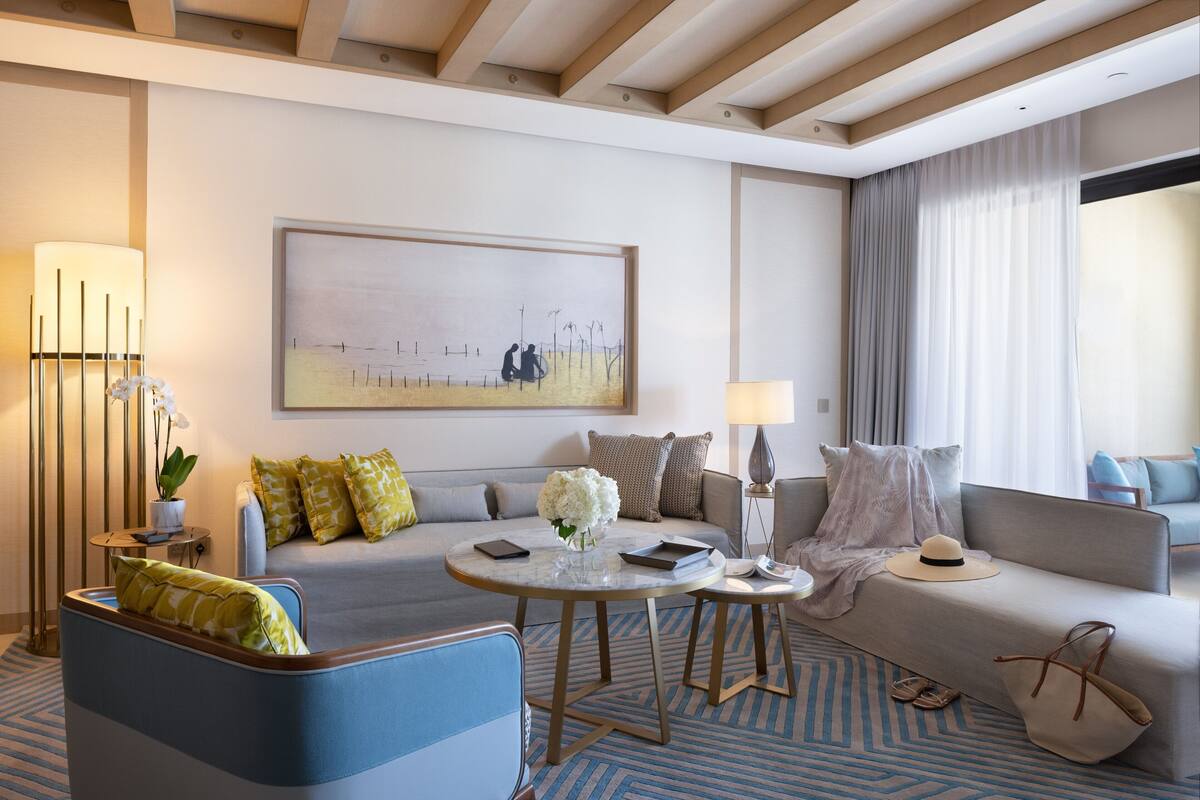A modern hotel room with a sofa, armchair, and a round coffee table. There is a painting on the wall, a lamp, and a window with curtains.