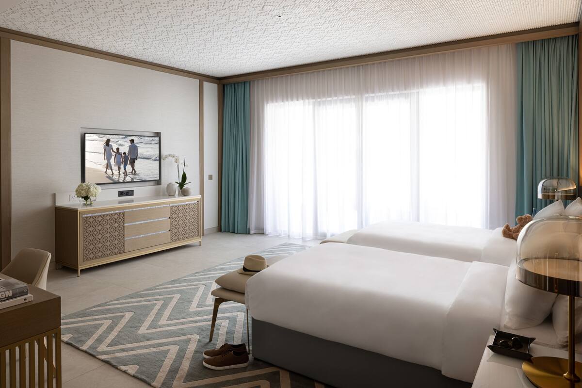 A hotel room with two beds, a TV, a desk, and a large window with curtains.