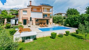 Una casa de dos pisos con piscina, mobiliario exterior y un porche cubierto.