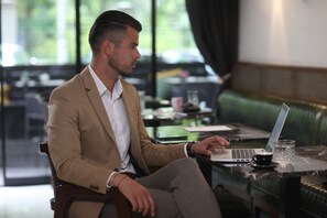 A man in a beige suit using a laptop at a table with a glass of water and a cup, in a room with a green upholstered sofa and a window with a view of trees. - Hotel Botanica (Belgrade)