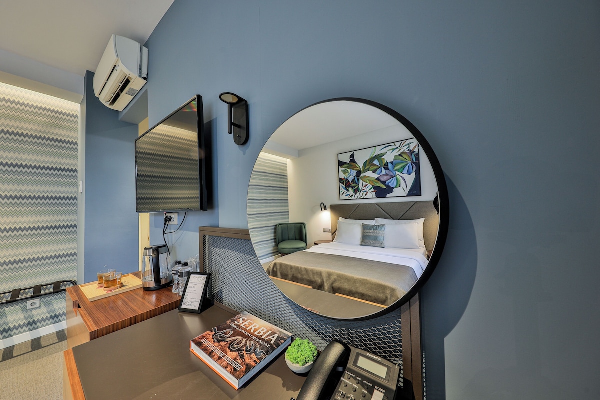 A hotel room with a bed, a round mirror, a desk with a phone, a coffee maker, and a framed book.