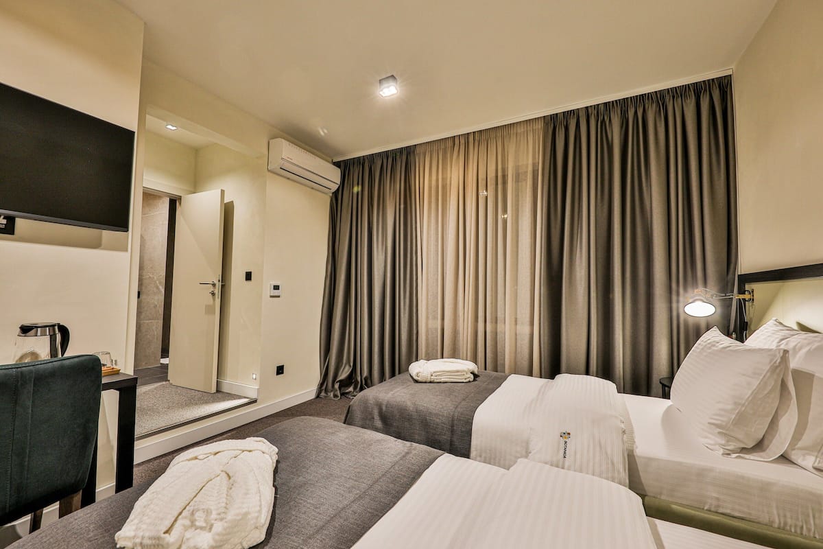 A hotel room with two beds, a desk, a chair, an air conditioning unit, and a large window with curtains.