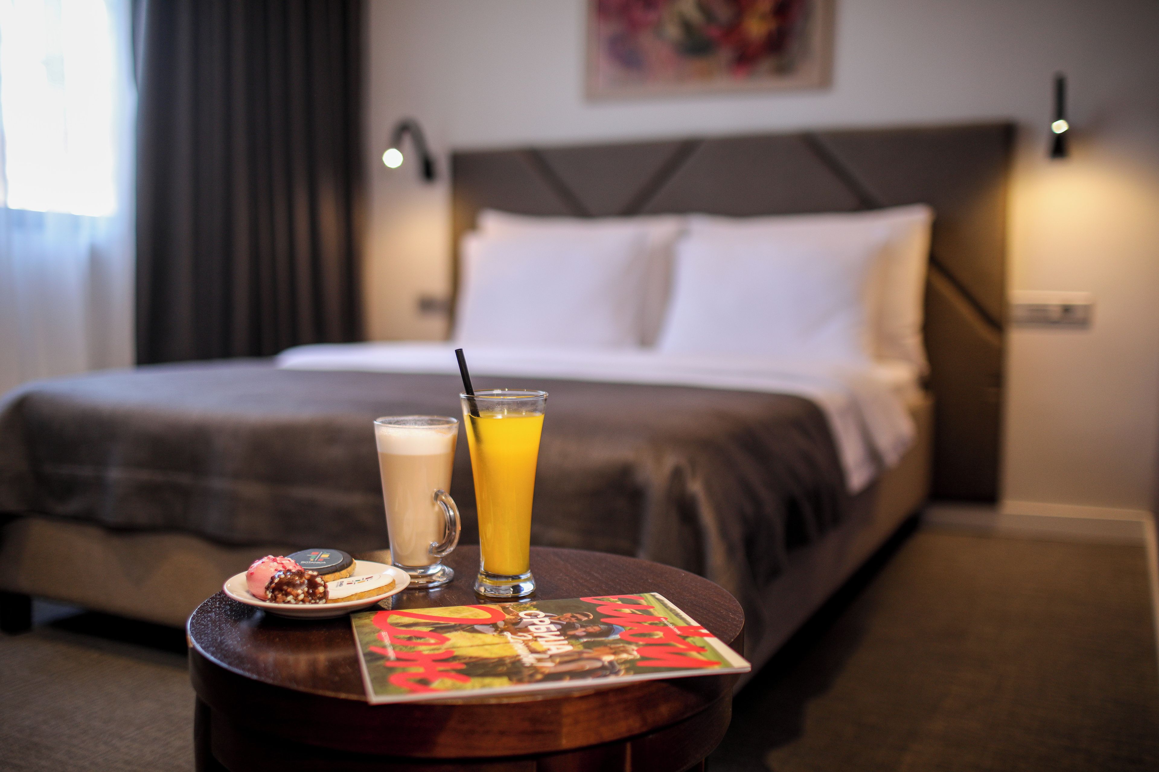 Une chambre d’hôtel avec un lit, une table sur laquelle se trouve un magazine, une assiette avec une pâtisserie et deux boissons.