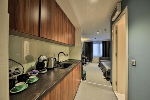 A compact kitchen with wooden cabinets, a coffee maker, and a sink. A small dining area with a table and chairs is visible in the background. - Hotel Botanica (Belgrade)