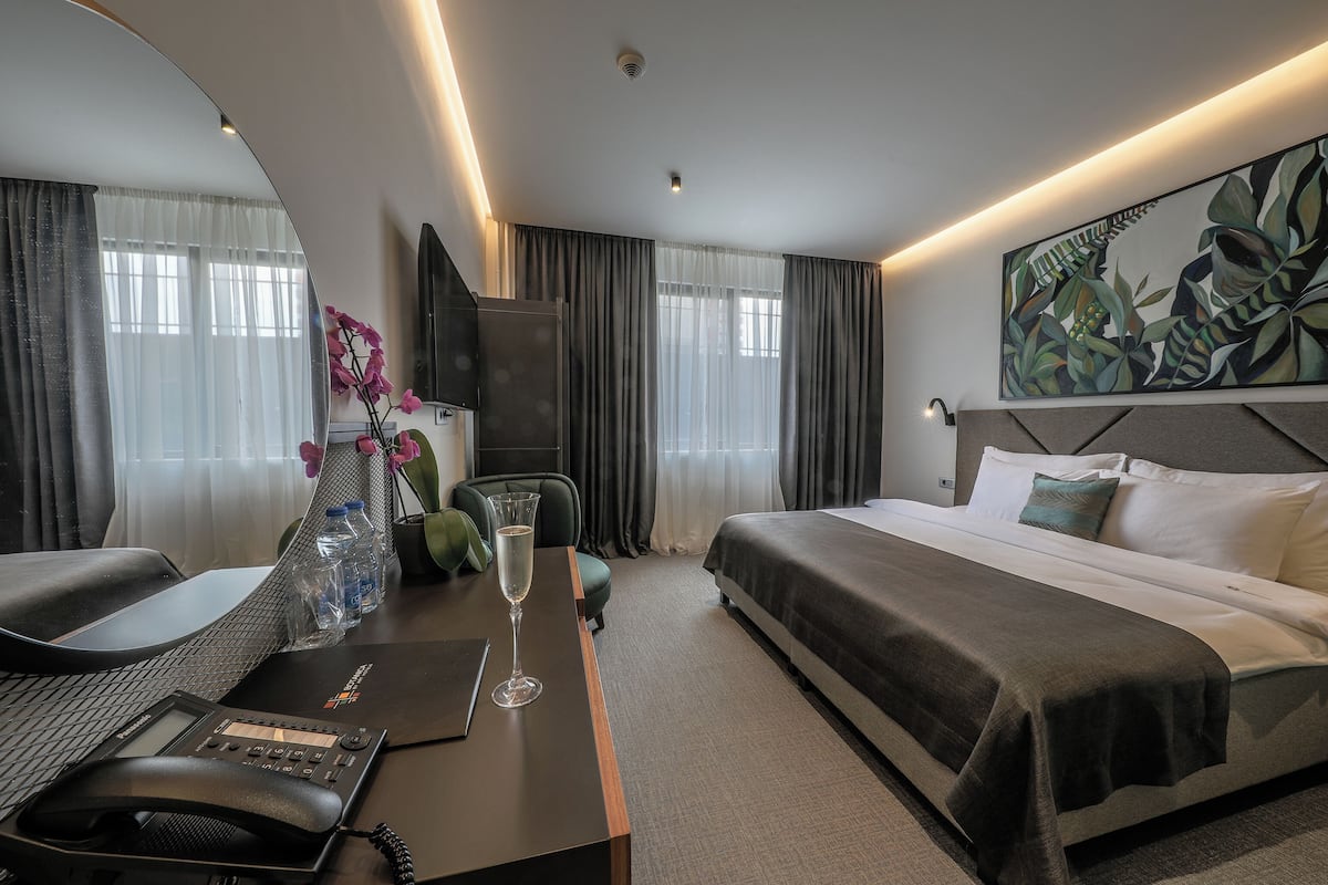 A hotel room with a bed, a desk with a phone, a chair, a glass of water, a vase with flowers, and a painting on the wall.