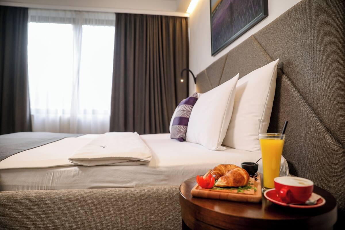 A hotel room with a neatly made bed, a breakfast tray with croissant, orange juice, and coffee, and a view of the city through large windows.