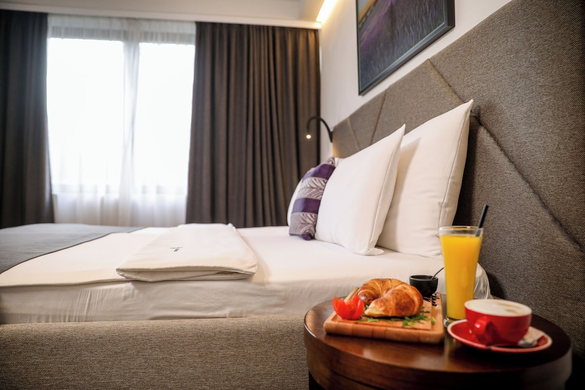 A hotel room with a neatly made bed, a breakfast tray with croissant, orange juice, and coffee, and a view of the city through large windows.
