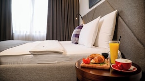 A hotel room with a neatly made bed, a breakfast tray with croissant, orange juice, and coffee, and a view of the city through large windows.