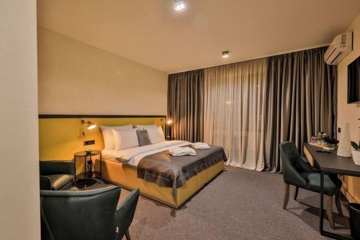 A hotel room with a bed, a desk, two chairs, and a large window with curtains.