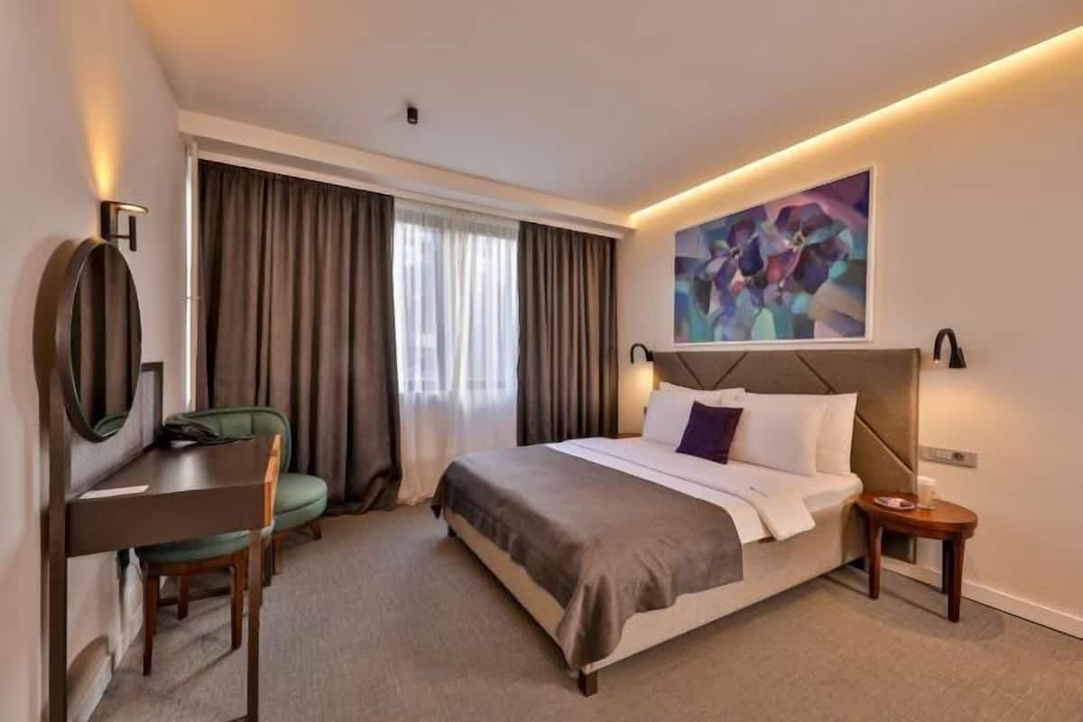 A hotel room with a bed, a desk, a chair, a small table, a painting on the wall, and a window with curtains.