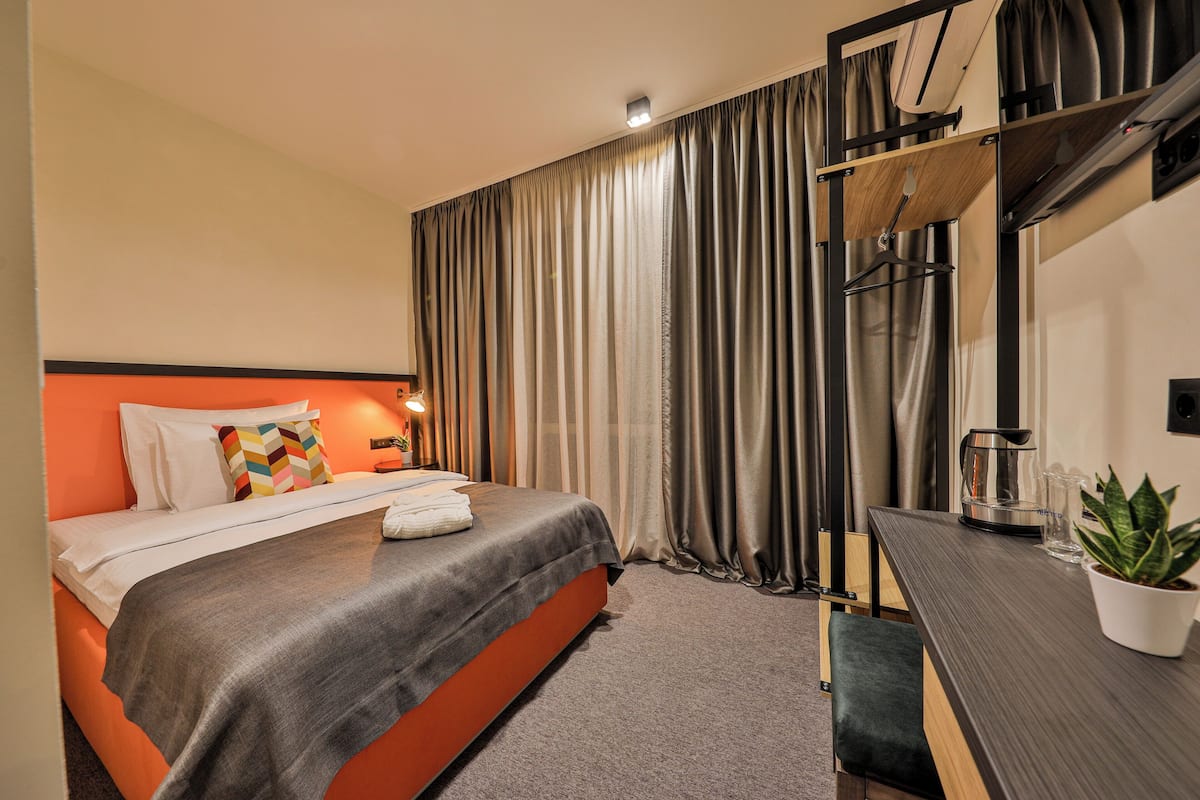 A hotel room with a bed, a desk, a potted plant, and a window with curtains.