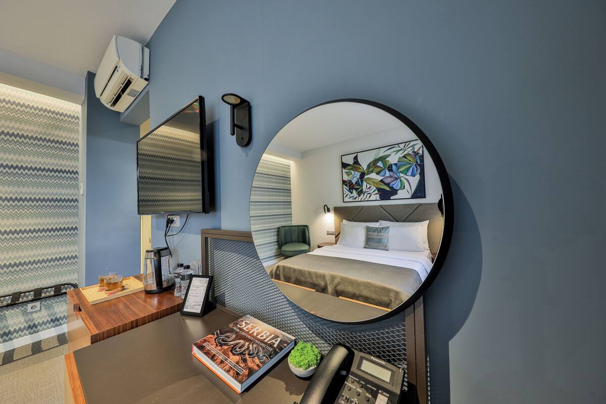 A hotel room with a bed, a round mirror, a desk with a phone, a coffee maker, and a framed book.