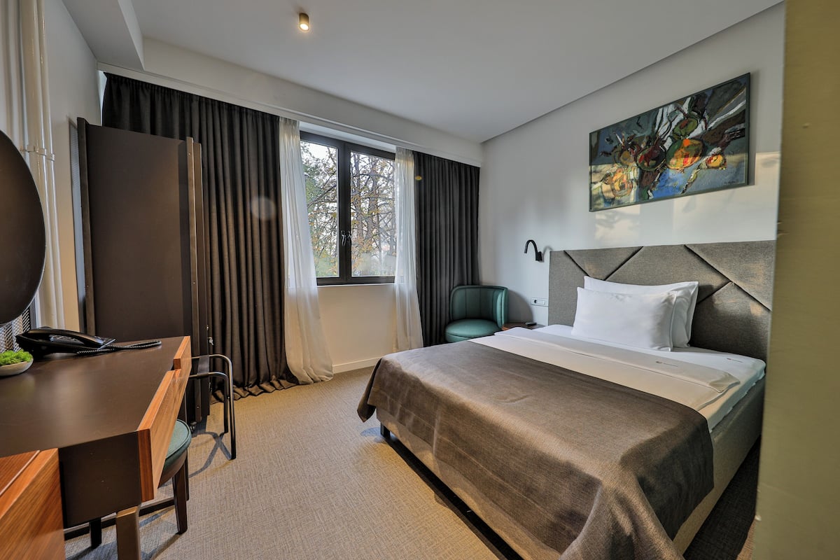 A modern hotel room with a large bed, a desk with a chair, a window with curtains, and a painting on the wall.