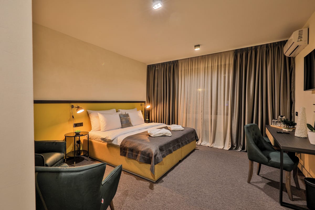 A hotel room with a bed, a desk, two chairs, a lamp, and an air conditioning unit.
