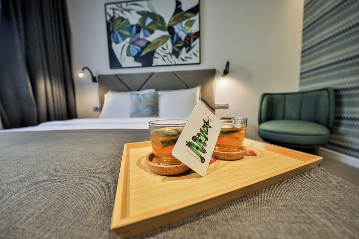 A hotel room with a bed, a bedside table with two glasses of amber liquid, a tray with a coaster and a card, a green chair, and a wall with a colorful abstract painting.