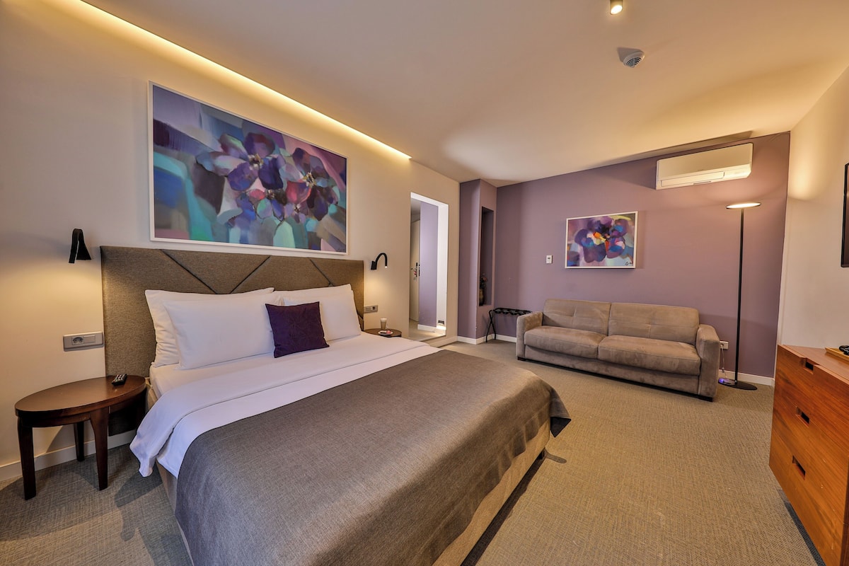 A modern hotel room with a large bed, a sofa, and two abstract paintings on the walls.