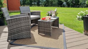 Eine Terrasse im Freien mit Korbmöbeln und einem für eine Mahlzeit gedeckten Tisch, mit Blick auf eine Rasenfläche und Bäume.