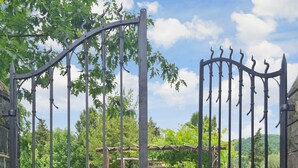 Un portail en fer forgé, doté d’un sommet incurvé et de barreaux verticaux, qui mène à un jardin agrémenté d’une pergola et d’un sentier.