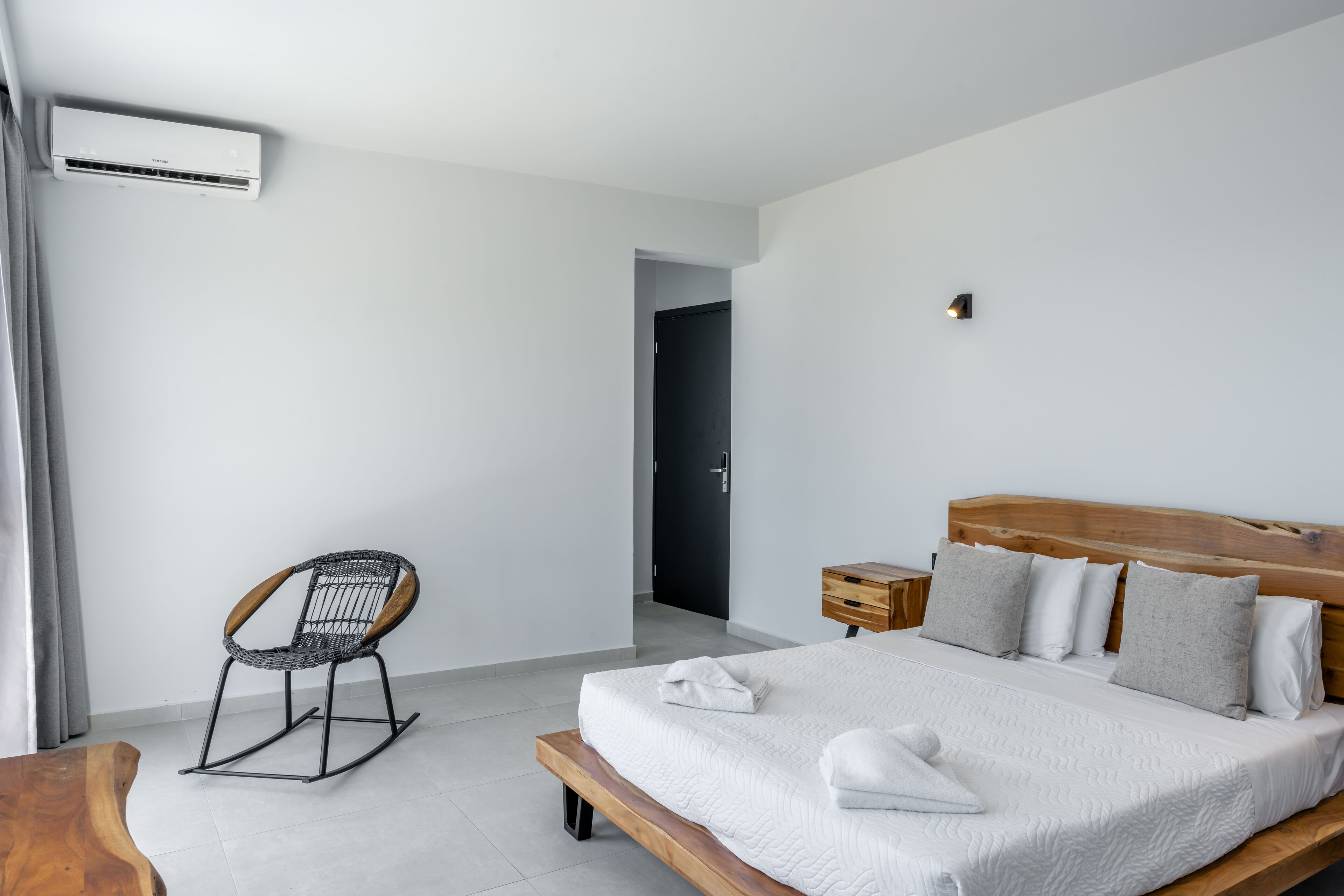 A modern hotel room with a wooden bed, a rocking chair, a bedside table, and a wall-mounted air conditioner.