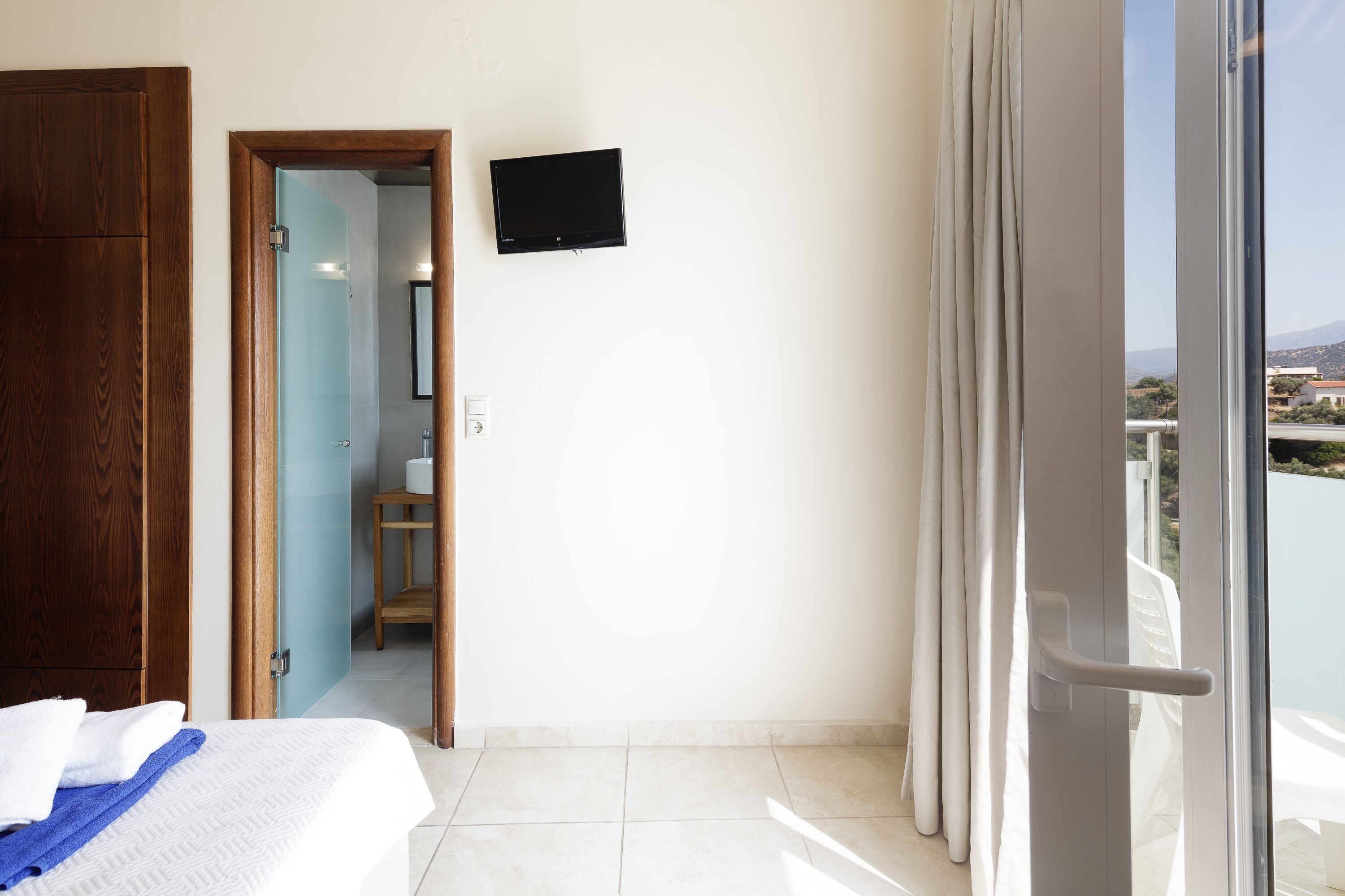 Une chambre d’hôtel avec un lit, une armoire en bois, un téléviseur fixé au mur et un balcon avec des rideaux.