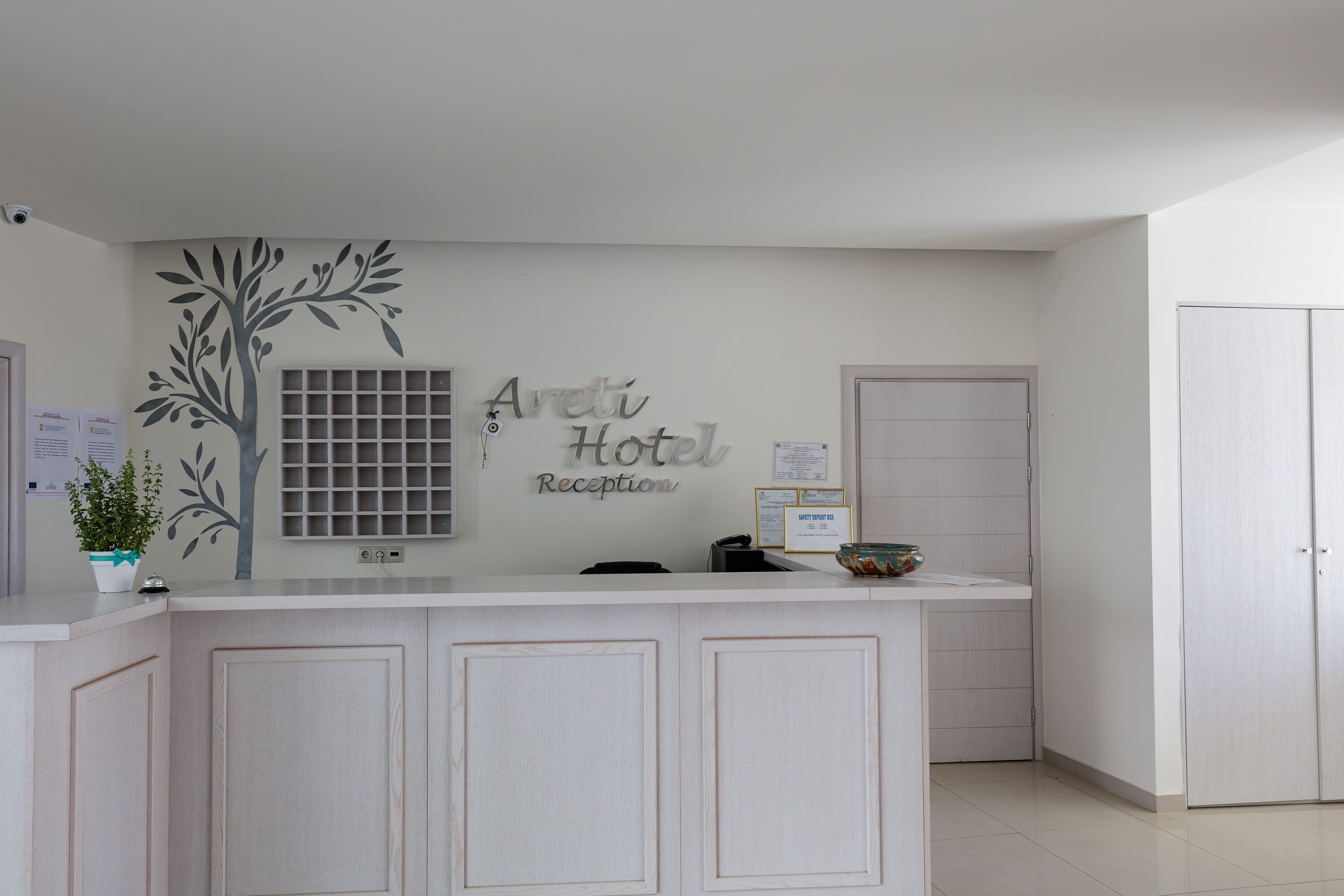 En receptionsdisk med skilt der viser "Areti Hotel Reception", en potteplante, et vindue med et gittermønster og en dør til højre.