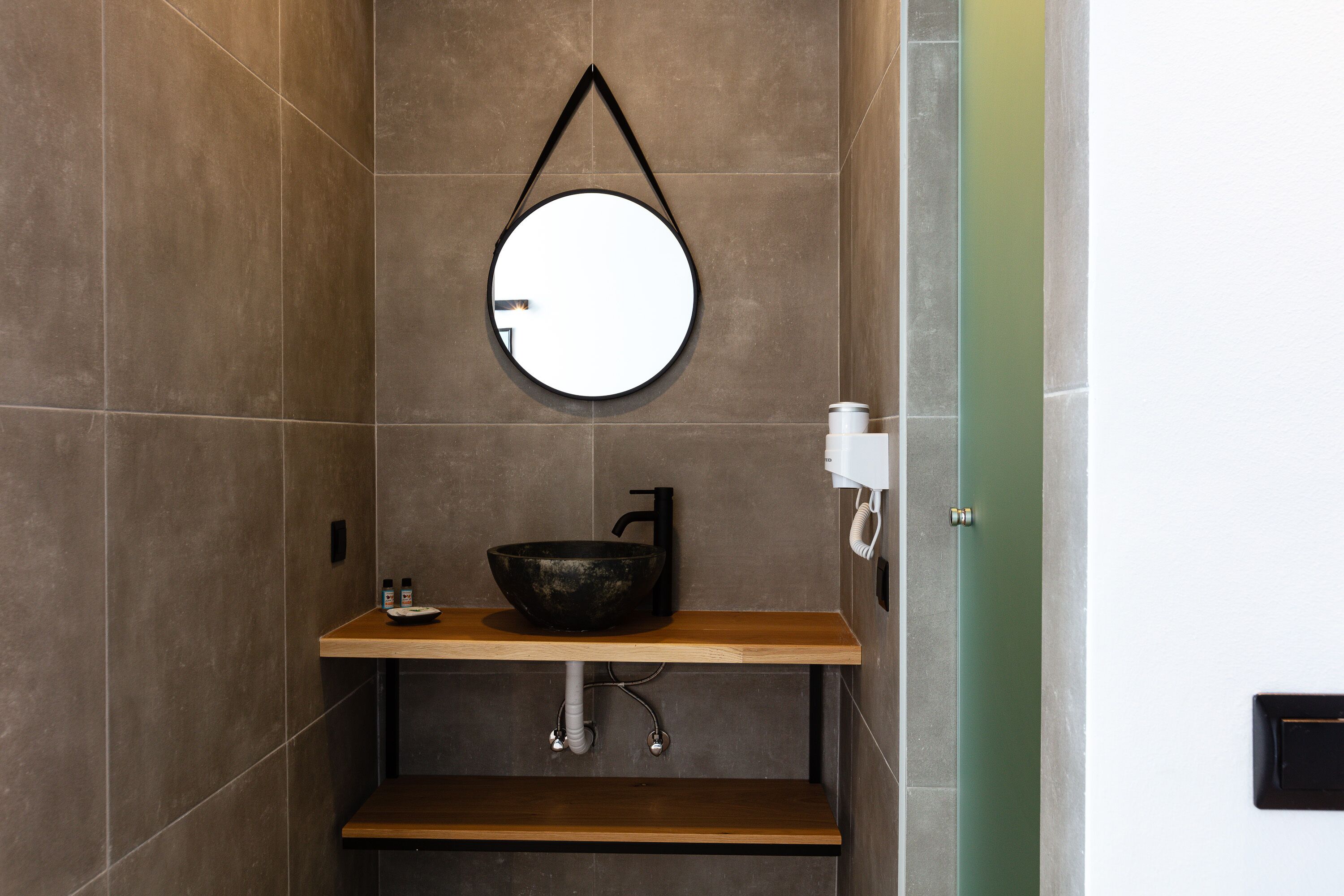 Une salle de bain avec un miroir rond, un lavabo noir et une étagère en bois.