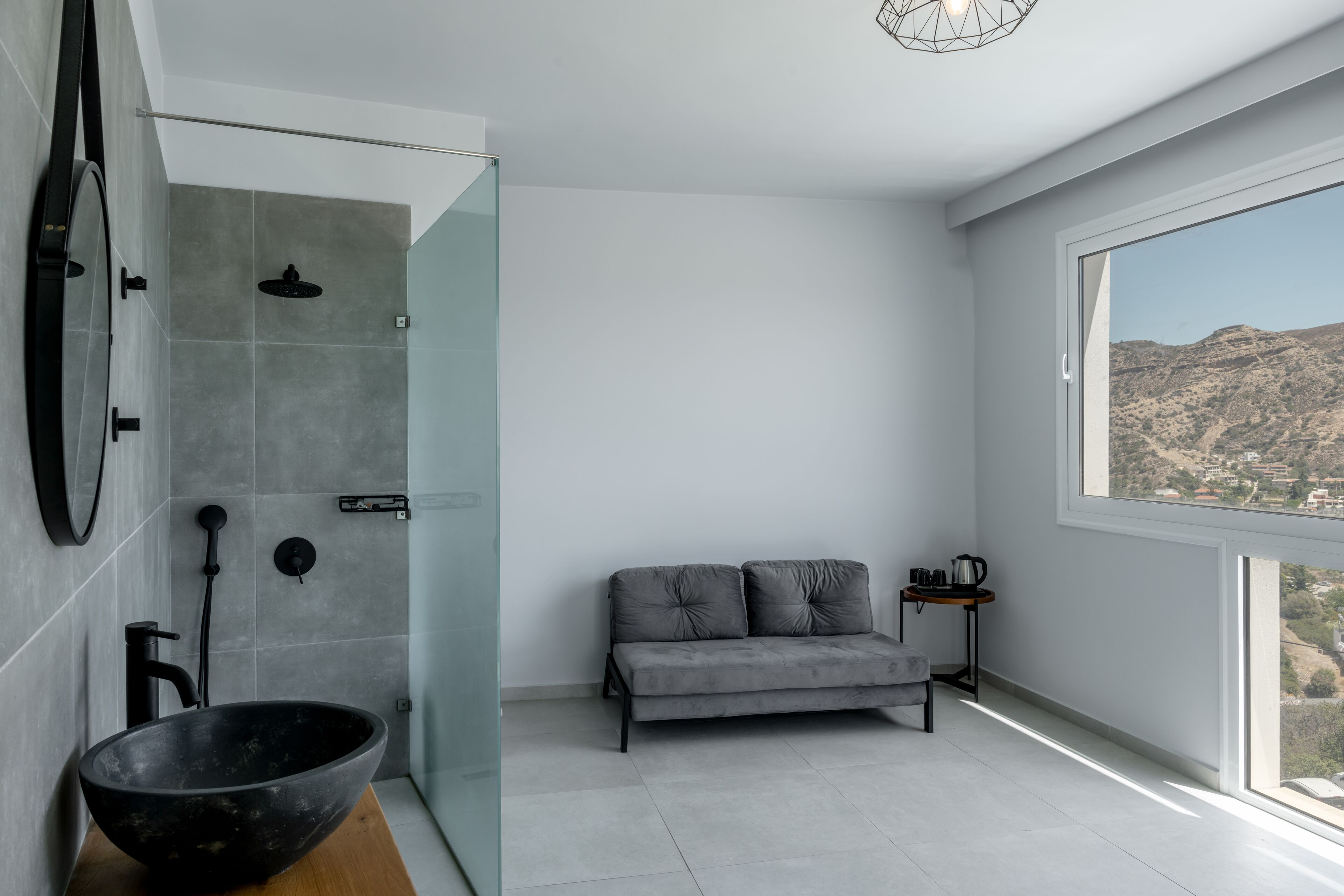 Ein modernes Badezimmer mit Duschkabine aus Glas, einem großen schwarzen Waschbecken und einem grauen Sofa an einem großen Fenster mit Blick auf die Berge.