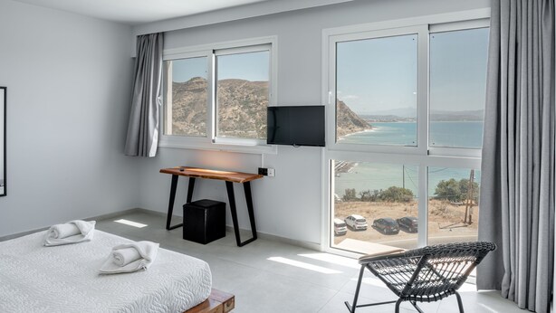 A modern hotel room with a large bed, a desk, a chair, and a TV. The room has large windows with a view of the sea and hills. - Areti Hotel (Agios Vasileios)