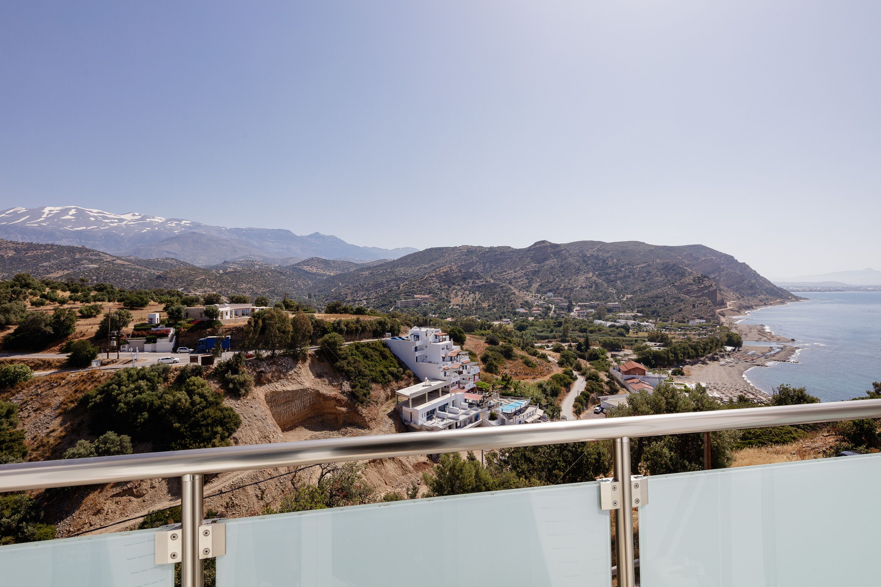 A coastal town with a mountain range in the background, a beach, and a residential area with houses and a swimming pool.