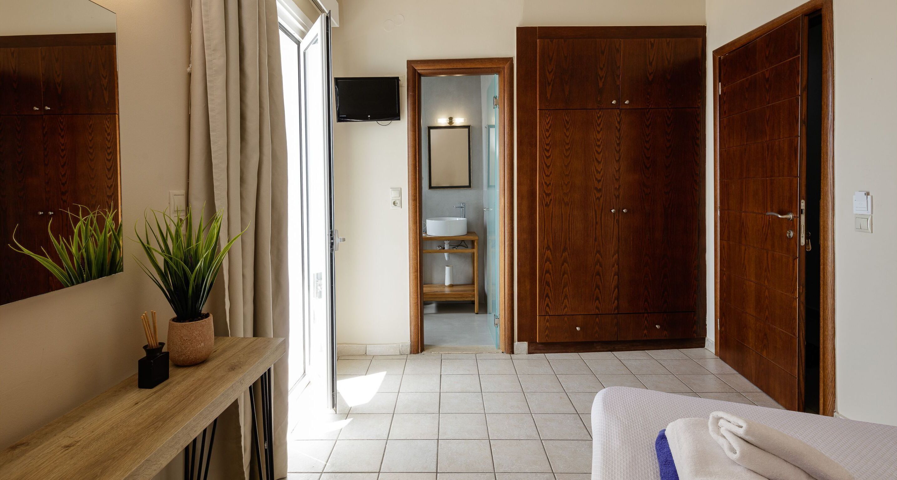 Una habitación de hotel con un armario de madera, una cama con manta blanca y azul, una consola de madera con una planta y un baño visible a través de una puerta abierta.