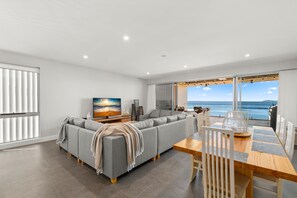A modern living room with a grey sofa, a wooden dining table, and a view of the sea through large windows.