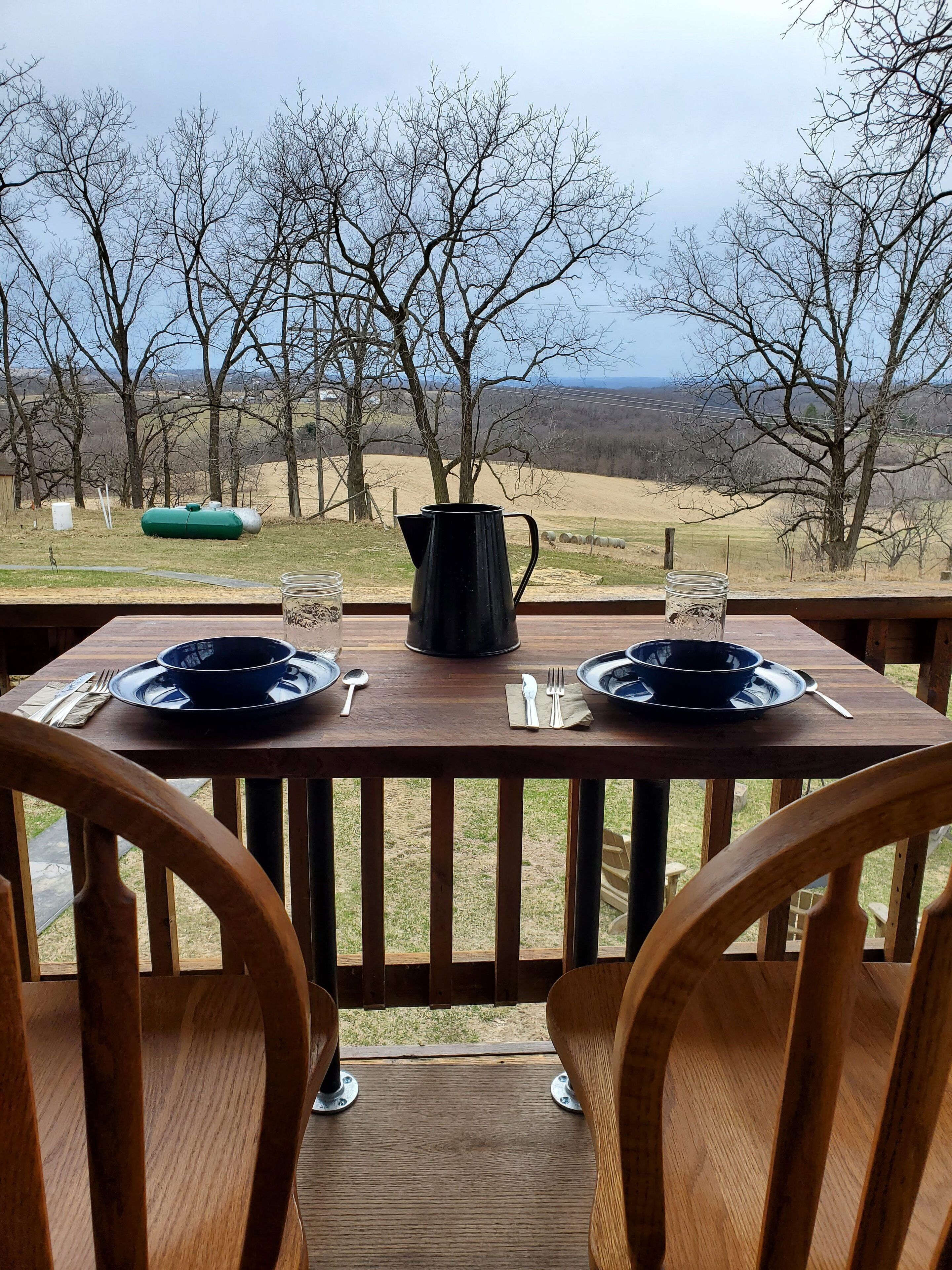 A wooden table set for two with a pitcher and glasses, overlooking a rural landscape with trees and a distant view.