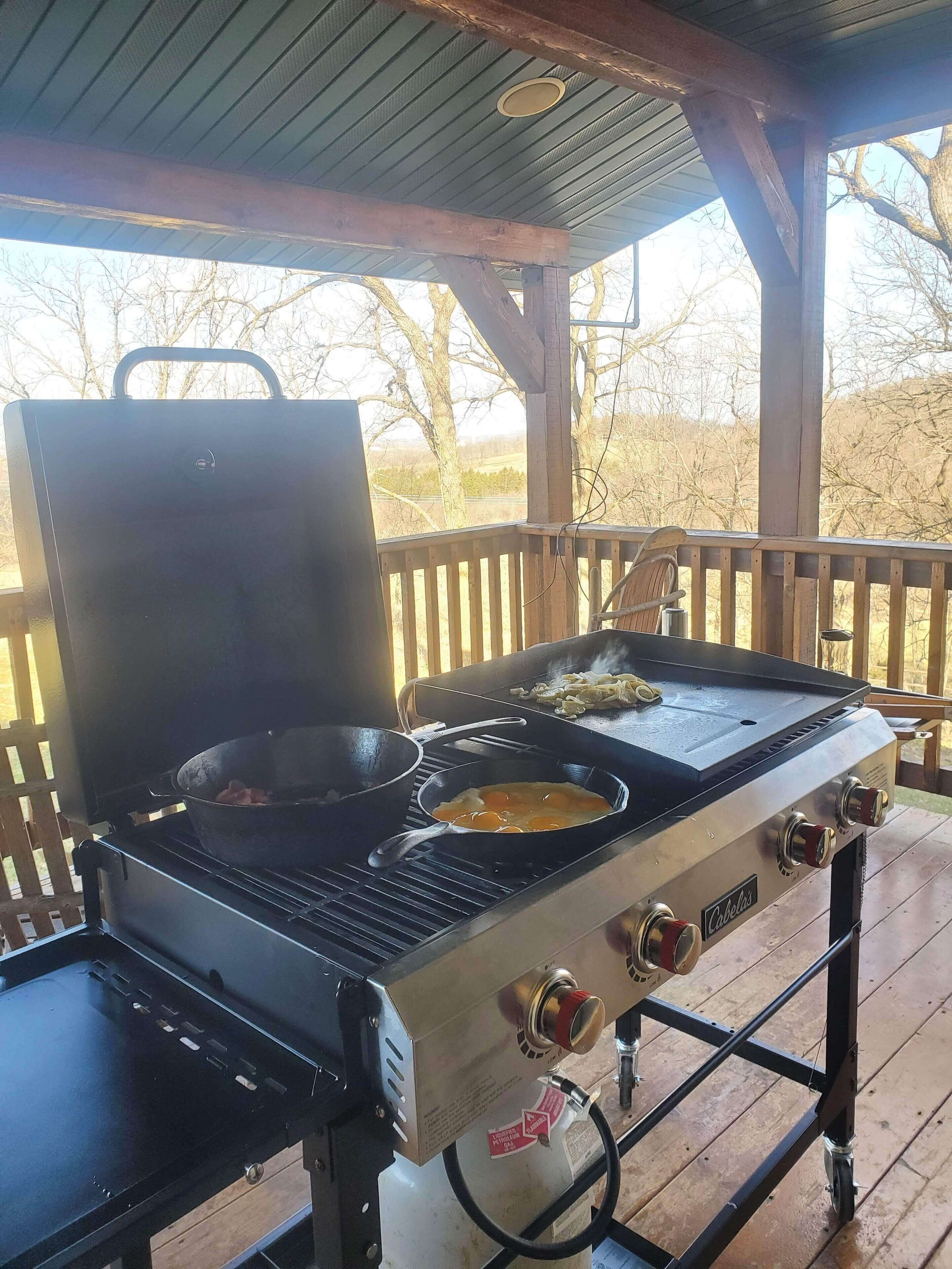 An outdoor cooking station with a grill and frying pans, set on a wooden deck with a view of trees and a clear sky.