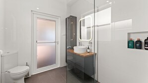 A modern bathroom with a toilet, a glass shower enclosure, a sink with a mirror, and toiletries on the wall. - Moorooba Pet Friendly Holiday Home Nelson Bay (Nelson Bay)