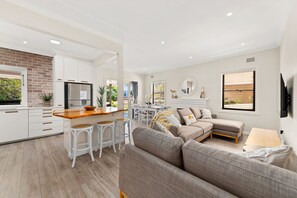 A modern kitchen with white cabinetry, a central island with stools, a dining area with a table and chairs, a living room with a sofa and a TV, and a window with blinds.