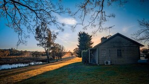 Een rustiek houten huisje met een kleine aangebouwd schuurtje, omringd door een grasveld en een waterpartij.