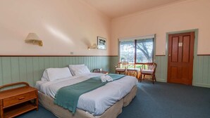 Un dormitorio ordenado con cama, mesita de noche de madera, silla, mesa y ventana con vistas a árboles.