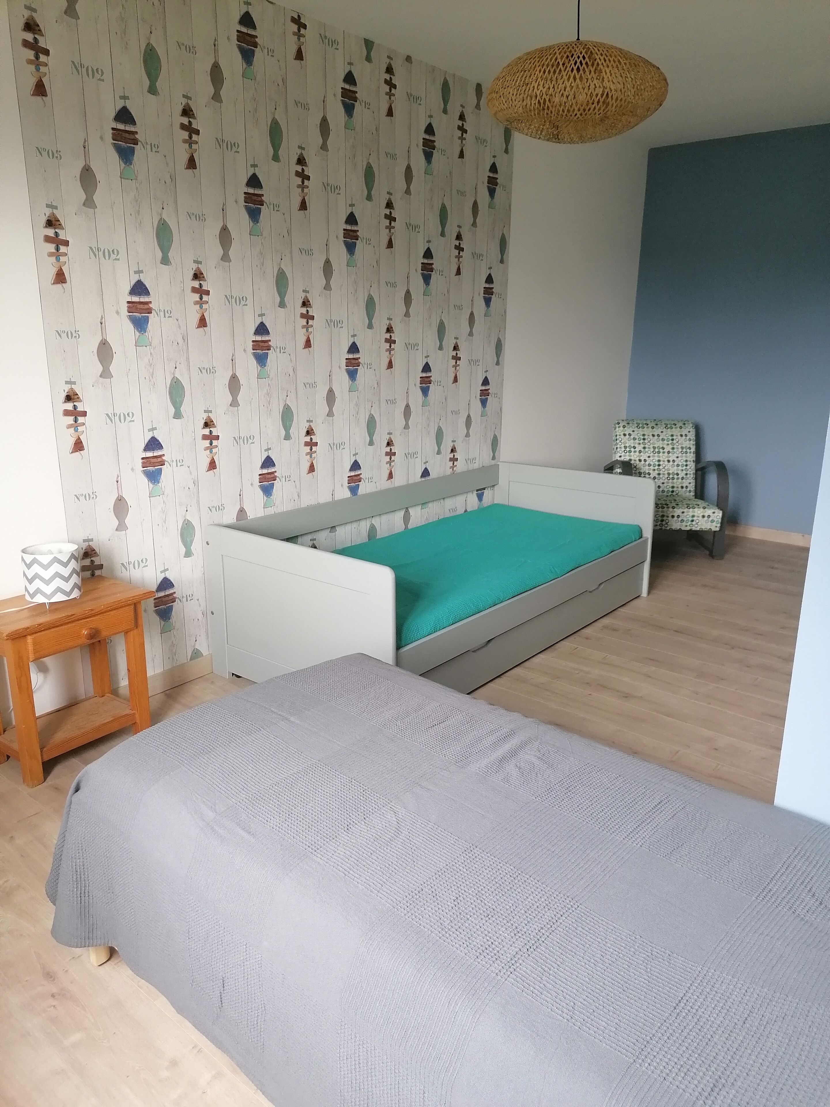 Een slaapkamer met een wit bed, een groene matras, een behang met patroon, een geweven lamp en een gestreept stoel.