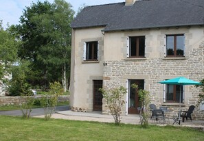 A two-story stone house with a stone facade, a dark roof, and a small front porch with outdoor seating. - Welcome to Brittany, where land meets sea. (Caulnes)