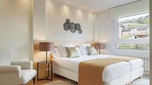 A modern bedroom with a large bed, two bedside lamps, a decorative wall piece, and a view of buildings through a large window. - Villa Loretopea by FeelFree Rentals (San Sebastián)