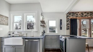 Una cocina moderna con gabinetes grises, electrodomésticos de acero inoxidable y ventanas amplias que dejan entrar luz natural.