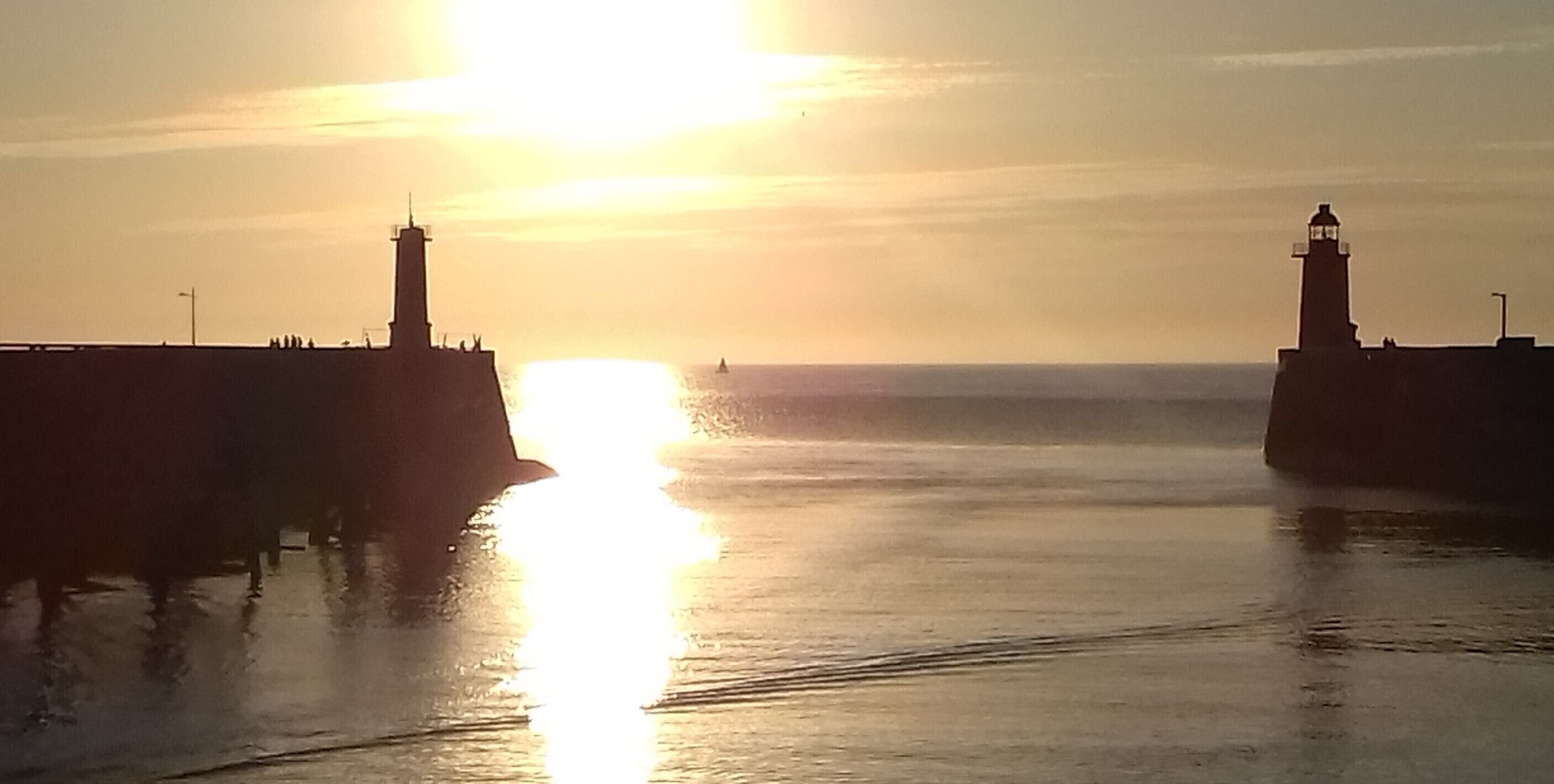 Ein Sonnenuntergang über ruhigem Wasser, mit zwei Leuchttürmen an gegenüberliegenden Seiten.