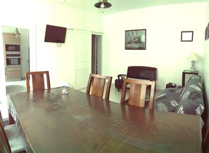 A dining area with a wooden table and chairs, a television mounted on the wall, and a kitchen in the background.